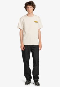 Camiseta beige de manga corta con un pequeño logo amarillo en el lado izquierdo del pecho, combinada con jeans de mezclilla oscura y zapatillas deportivas negras.