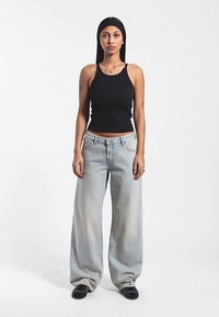 Camiseta negra sin mangas con textura acanalada, vaqueros de pierna ancha azul claro con lavado descolorido y zapatos negros. Modelo posando frente a un fondo blanco liso.