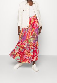 Person in einer weißen Jacke, einem weißen Oberteil, einem bunten floralem Maxi-Rock und weißen Sneakers, der vor einem schlichten hellen Hintergrund steht.