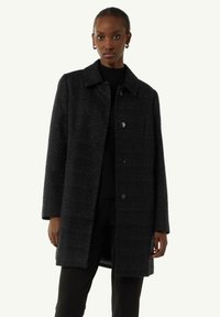 Black textured coat with a collar, button closures, and long sleeves. Worn over a black top, features a straight cut and minimal design.