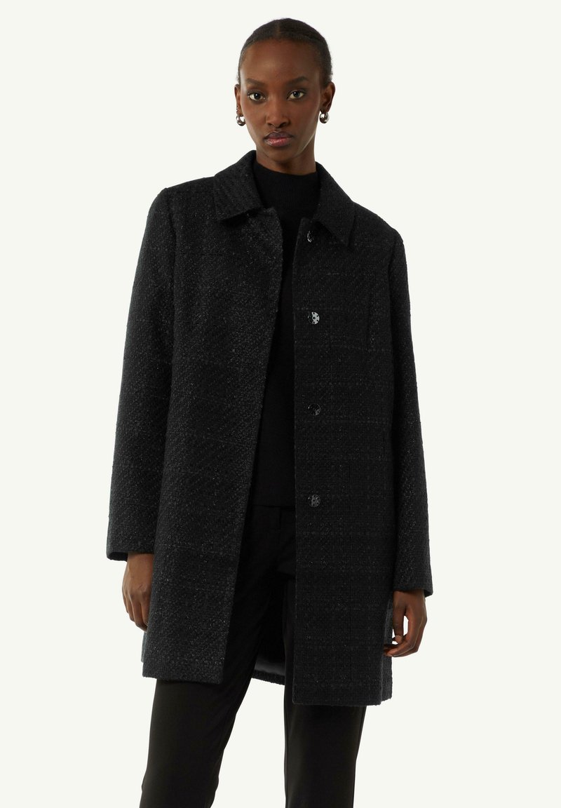 Black textured coat with a collar, button closures, and long sleeves. Worn over a black top, features a straight cut and minimal design.