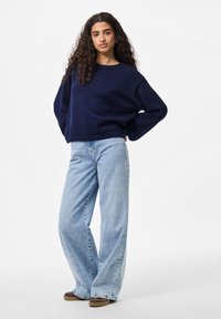 Suéter tricotado azul-marinho com um corte descontraído e mangas largas, combinado com jeans de perna larga azul-claro e sapatos castanhos. Fundo simples.