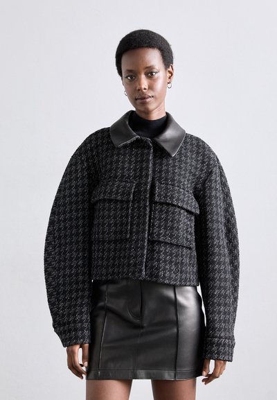 Veste noire texturée court avec de grandes poches avant et un col noir brillant, associée à une jupe en cuir noir ajustée.