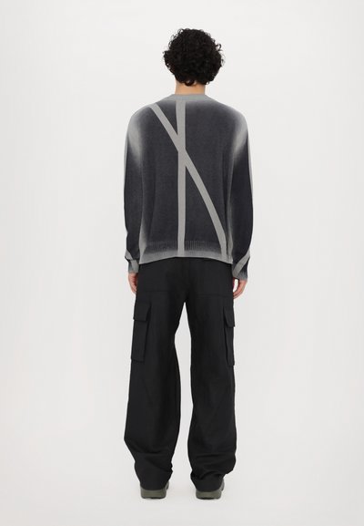 Gestrickter grauer Pullover mit einem helleren grauen diagonalen Streifendetail, getragen mit lockeren schwarzen Cargo-Hosen mit mehreren Taschen.