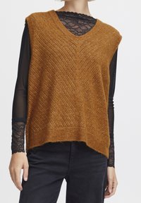 Miedziany sweter bez rękawów o splotach z dekoltem w szpic, zdobiony wzorem jodełki i fakturą, noszony na czarnym koronkowym topie z długim rękawem.