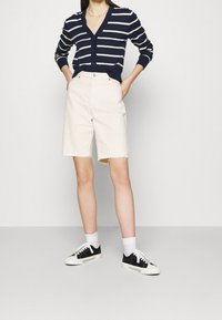Cárdigan de rayas navy con botones, combinado con pantalones cortos de mezclilla crema con bordes deshilachados. El modelo lleva zapatillas negras y calcetines blancos.