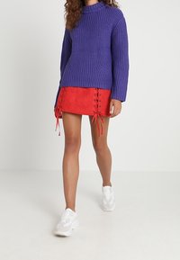 Fialový pletený svetr s vysokým výstřihem, doplněný červenou semišovou mini sukní s bočním šněrováním. Bílé sportovní boty.