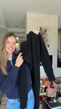 Smilende kvinde i blå sweater holder en sort langærmet skjorte op inde i en moderne stue med kunst og dekorationer.