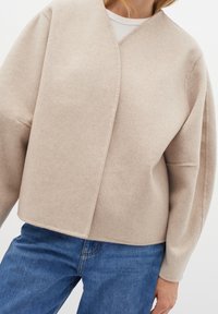 Bežinės vilnos mišinio apykaklės megztinis su minkšta tekstūra, giliu V formos iškirpimu ir laisvomis rankovėmis, derinamas su mėlynomis džinsinėmis kelnėmis.