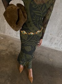 Dlhé rukávy, priliehavé šaty v tmavozelenej farbe s kvetinovými vzormi, doplnené zlatým opaskom s oválnymi zelenými kameňmi. Hnedá semišová spojka a béžové podpätky so cvočkami.