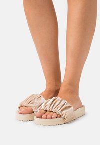 Velvet slide sandals with ruched upper, beige color, and textured sole for grip. Open-toe design highlighting toes.