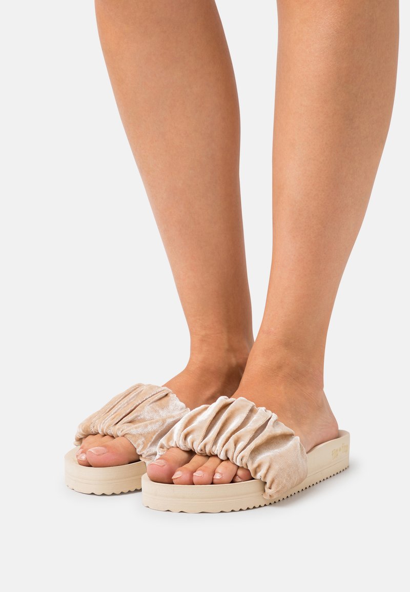 Velvet slide sandals with ruched upper, beige color, and textured sole for grip. Open-toe design highlighting toes.