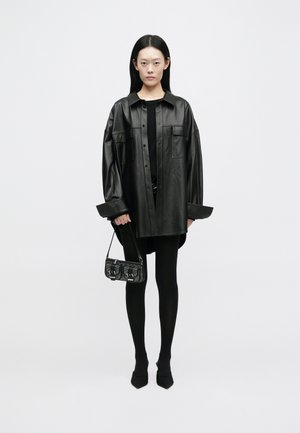 Camicia giacca oversize in pelle nera con tasche frontali, abbinata a un top nero aderente, collant neri e una piccola borsa testurizzata.