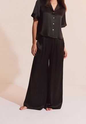 Femme portant un haut noir en satin à manches courtes, ample, boutonné, et un pantalon à jambes larges, debout pieds nus devant un fond uni.