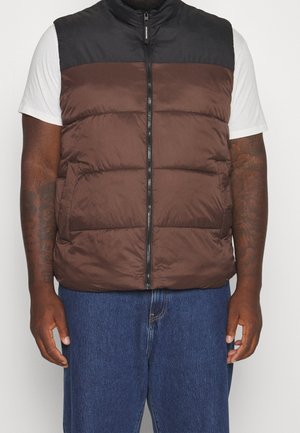 Homme portant une veste matelassée sans manches marron et noire sur une chemise blanche à manches courtes et un jean bleu, debout les bras détendus.