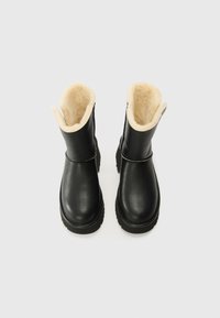 Botine negre din piele, cu căptușeală din blană bej vizibilă pe vârful pliat, talpă groasă și vârf rotund, pe fundal alb.