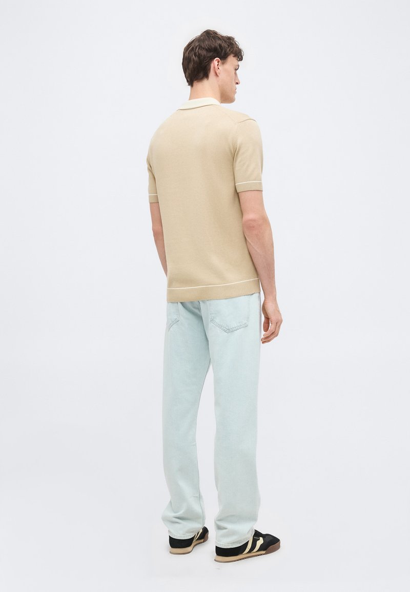 Homme portant un polo à manches courtes beige, un jean bleu clair et des baskets noires à rayures blanches, debout et tourné de dos sur un fond uni.