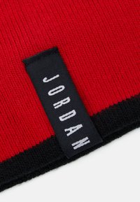 Bonnet en tissu côtelé rouge avec une étiquette noire portant "JORDAN" en texte blanc, avec un bord inférieur noir contrasté.