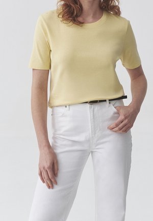 Osoba nosząca bladożółtą bluzkę z krótkim rękawem wpuszczoną w wysokie białe spodnie z czarnym paskiem, ręka spoczywa blisko kieszeni.