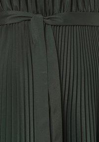 Faltenstoff in Dunkelgrün mit glatter Textur, präsentiert mit einem passenden Gürtel, der in der Taille gebunden ist und das strukturierte Design des Kleidungsstücks betont.