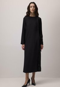 Vestido negro de manga larga con corte recto, que presenta una manga izquierda adornada con lentejuelas y una textura de tela suave. Combinado con tacones negros.