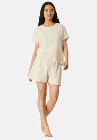 Frau steht barfuß und trägt ein cremefarbenes Kurzarmshirt sowie passende Shorts mit elastischem Bund vor neutralem Hintergrund.