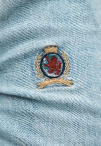 Κεντημένο έμβλημα σε γαλάζιο τζιν που απεικονίζει ένα κόκκινο λιοντάρι μέσα σε κυκλικό πλαίσιο με χρυσή κορυφή και στεφάνι δάφνης.