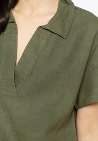 Blusa de manga corta en verde oliva con escote en V y cuello, hecha de una tela ligera que presenta una superficie suave y ligeramente texturizada.