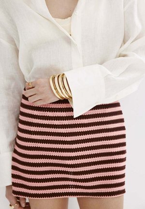 Femme portant un chemisier blanc avec de larges poignets et une jupe en maille rayée rose et marron, accessoirisée de plusieurs bracelets et bagues en or.