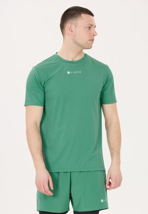 Mand iført grøn Virtus sport T-shirt og matchende shorts, stående med afslappede arme, der kigger til siden mod en ensfarvet baggrund.