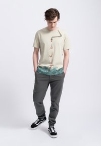 Beige t-shirt met korte mouwen met een afbeelding van een waterpijp en figuren, gecombineerd met donkergrijze sweatpants en zwarte sneakers.