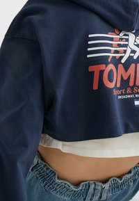 Sudadera recortada azul marino con texto gráfico en blanco y rojo, superpuesta a una camiseta blanca, combinada con pantalones cortos de mezclilla de tiro alto, mostrando un borde deshilachado.