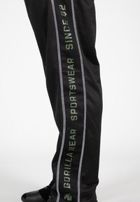Zwarte sportbroek met groene tekst "GORILLA WEAR" en "SPORTSWEAR SINCE 82", voorzien van zilveren strepen en een mesh-textuur.