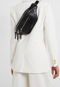 Borsa a tracolla in pelle nera con scomparti zip multipli, design elegante, texture liscia e cinturino regolabile, indossata su un abbigliamento bianco.