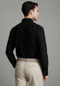 Cămașă neagră cu mâneci lungi și guler, prezentată din spate. Asortată cu pantaloni bej deschis și o curea texturată împletită. Material neted.