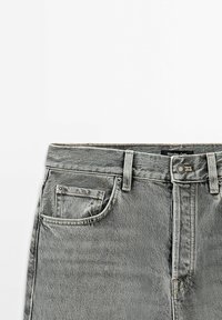 Détail de la taille d’un jean en denim gris avec bouton, passants de ceinture, poche avant et étiquette Massimo Dutti sur fond blanc.