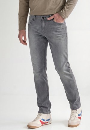 Homme portant un jean slim gris, une chemise beige à manches longues et des baskets blanches avec des rayures bleues et rouges, debout avec une main dans la poche.