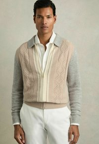 Cardigan tricotat în bej și gri cu un tiv cu nervuri, modele texturate în formă de diamant, închidere cu fermoar în față și guler contrastant peste o cămașă albă.