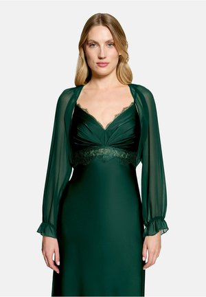Frau mit langen blonden Haaren, die ein dunkelgrünes Kleid mit transparenten langen Ärmeln und Spitzenbesatz am Oberteil trägt, vor einem einfarbigen Hintergrund steht.