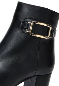 Bottine en cuir noir avec un bout carré, un épais talon carré et un accent de boucle rectangulaire dorée sur le côté. Fini texturé lisse.