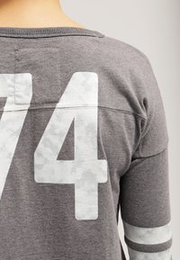 Grå långärmad tröja med rund halsringning, som har ett stort vitt nummer "74" och vita randiga detaljer på ärmarna. Mjuk tygtextur.