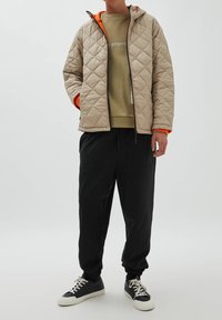 Veste matelassée beige avec une capuche orange, portée sur un sweat-shirt vert, associée à un pantalon de jogging noir et des baskets noires.