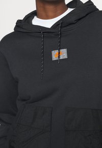 Svart huva sweatshirt med kontrasterande texturerad svart ficka, grå logotypflik och reflekterande dragsnören. Blanda bomullstyg.