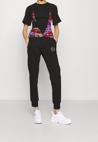 Svart t-shirt med kort ärm och flerfärgad mönstrad panel, tillsammans med svarta joggers med en logotyp-patch och vita sneakers.