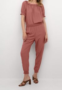 Roodachtig roze jumpsuit met een rimpelnek, korte mouwen en een elastische tailleband. Beschikt over taps toelopende pijpen en verzamelde manchetten. Bruine haksandalen.