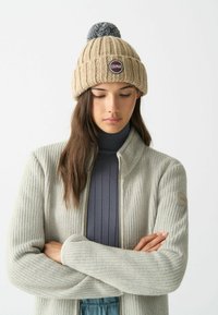 Berretto lavorato a maglia beige con un pom-pom grigio e una toppa rotonda del marchio. Il modello indossa un cardigan grigio chiaro con zip, in tessuto testurizzato, sopra una maglia scura.