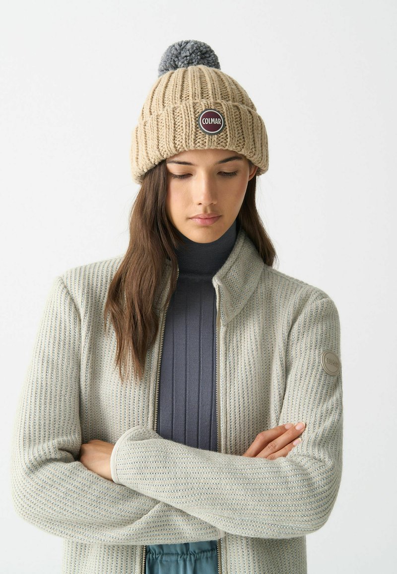 Berretto lavorato a maglia beige con un pom-pom grigio e una toppa rotonda del marchio. Il modello indossa un cardigan grigio chiaro con zip, in tessuto testurizzato, sopra una maglia scura.