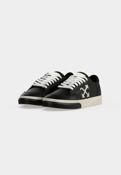 Ζευγάρι μαύρα sneakers χαμηλού ύψους με λευκά κορδόνια, λευκές λαστιχένιες σόλες και λευκό λογότυπο σταυρωμένου βέλους στο πλάι.