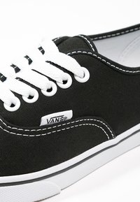 Baskets en toile noire avec lacets blancs et surpiqûres blanches, dotées d'une semelle en caoutchouc blanche et d'une petite étiquette de marque sur le côté.