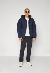 Mörkblå vadderad jacka med huva, dragkedja framtill och sidofickor, bärs över en vit skjorta och kombineras med svarta byxor och sneakers.